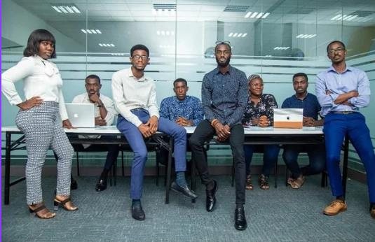 Seven members of the team posed on and around a conference table in a glass-walled office